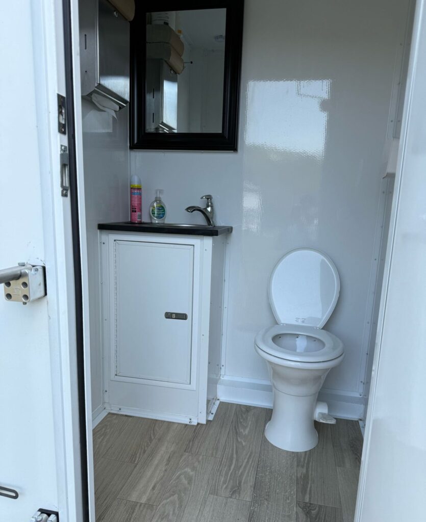 Private bathroom trailer stall with a flushing toilet, vanity sink, framed mirror and wood-look floor, The Lavatory Los Angeles