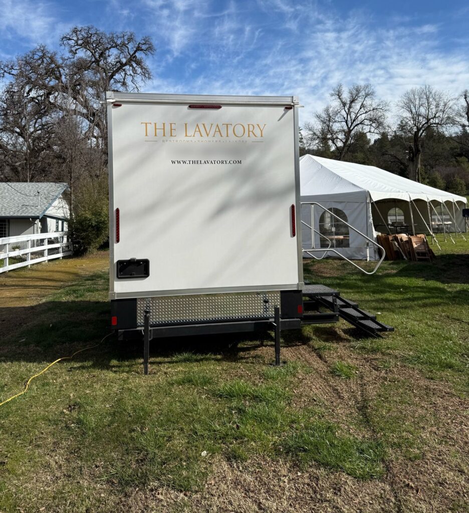 Luxury bathroom trailer with entry steps set on a grass lawn next to a white wedding tent and stacked wood chairs, The Lavatory Los Angeles