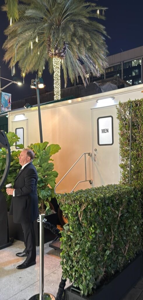 Event staff member beside a hedge-screened 5-station bathroom trailer at a nighttime store opening in Beverly Hills, The Lavatory Los Angeles