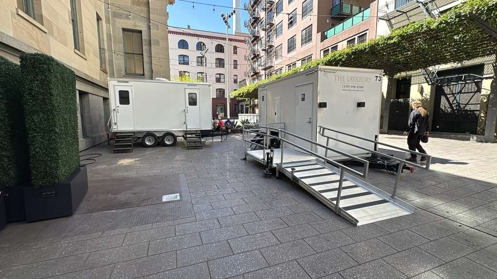 Accessible bathroom trailer with a railed ramp set beside a standard bathroom trailer in a brick city courtyard, The Lavatory Los Angeles