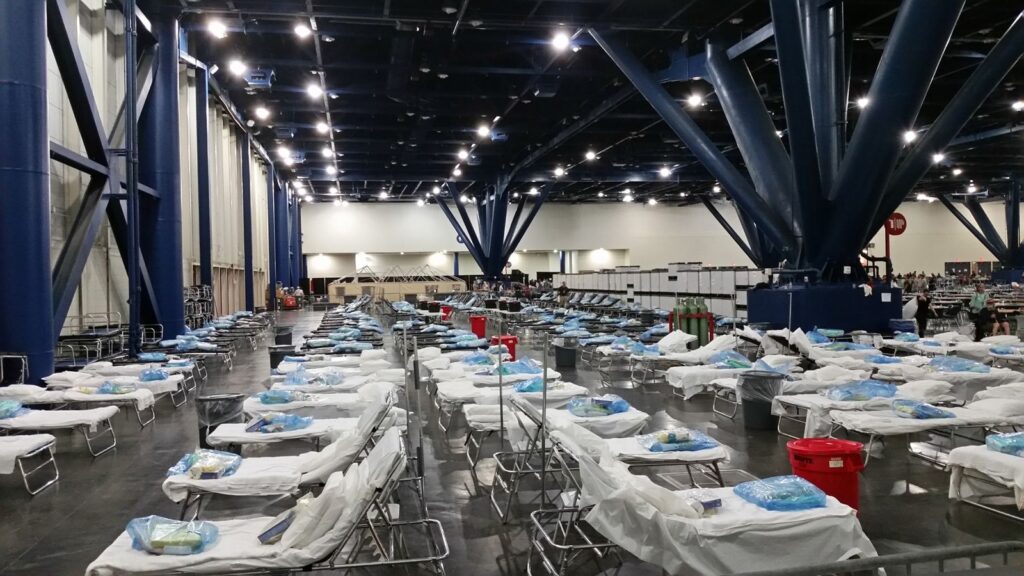 Rows of cots with blankets set up inside a large evacuation shelter hall, The Lavatory Los Angeles