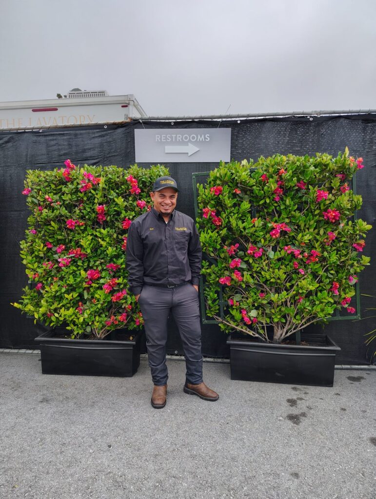 Smiling restroom attendant from our team standing between flowering hedge walls under a restrooms sign, The Lavatory Los Angeles