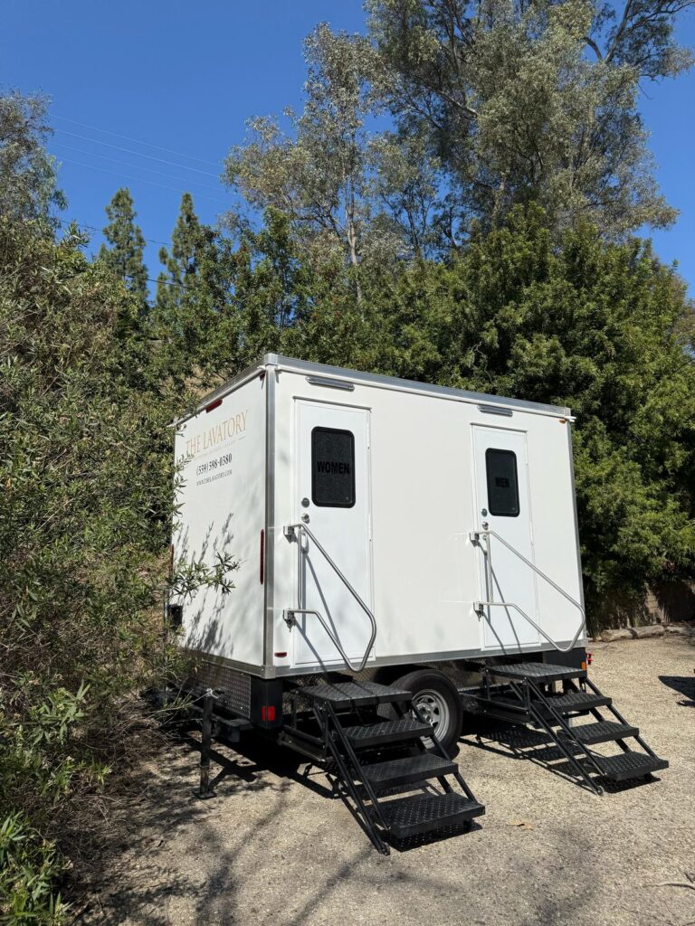 2-station bathroom trailer with women's and men's doors and steps on a shaded gravel lot in Fullerton, The Lavatory Los Angeles