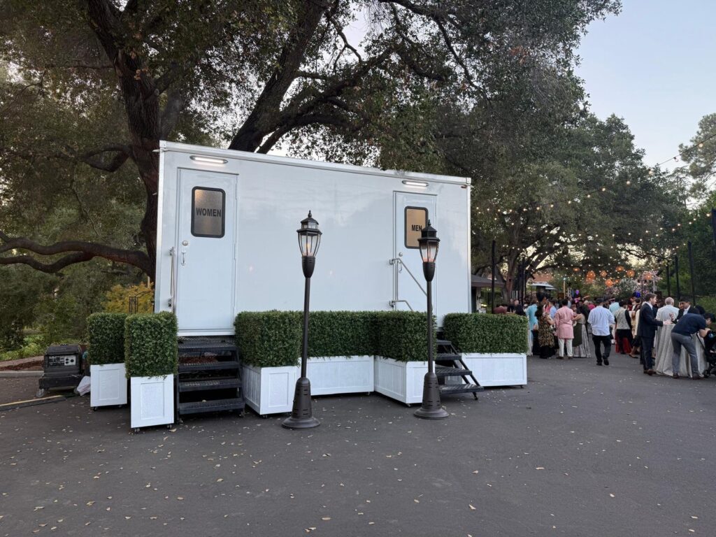 Guests at an evening gala under oak trees and string lights beside a hedge-screened bathroom trailer with lamp posts, The Lavatory Los Angeles