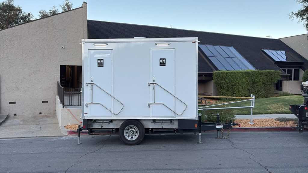 Compact 2-station bathroom trailer parked at the curb outside a school building with solar panels on the roof, The Lavatory Los Angeles