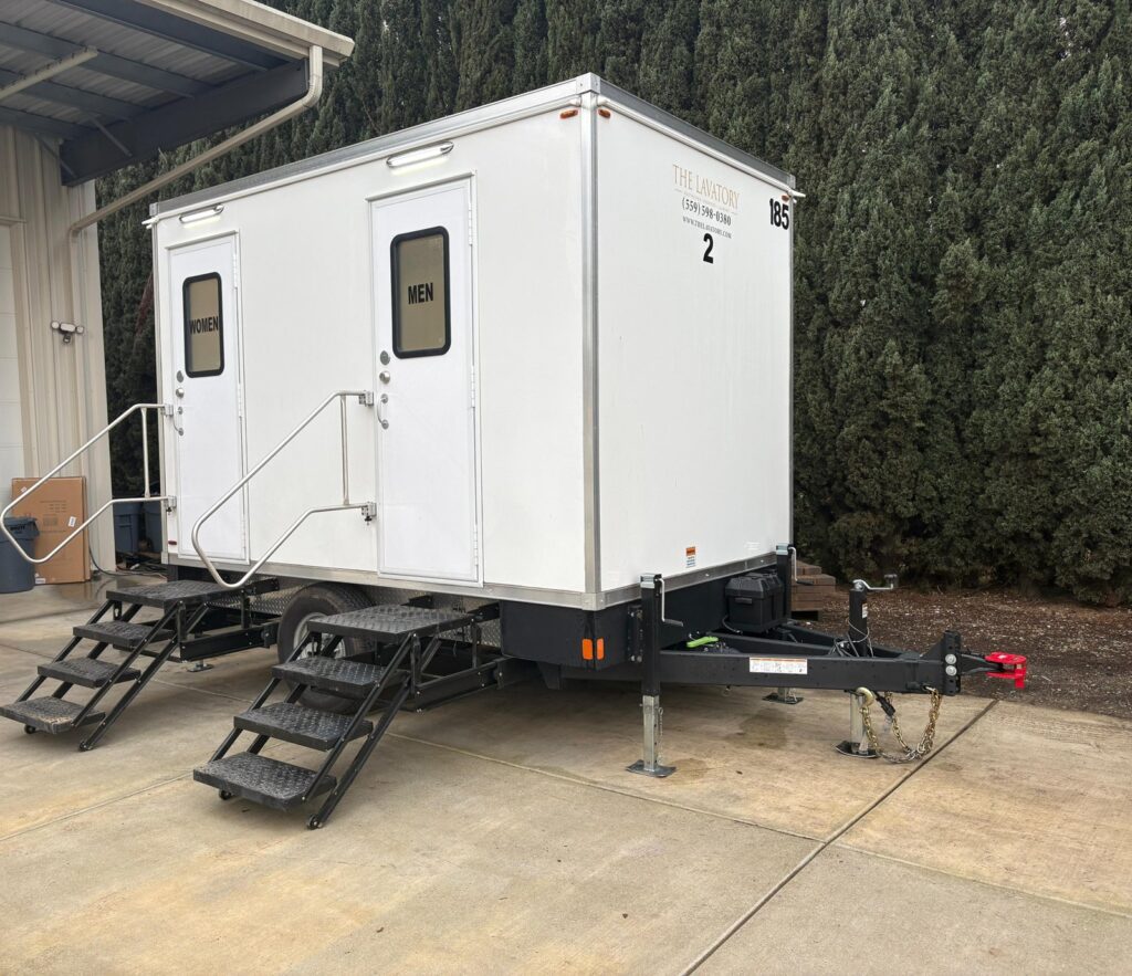 2-station bathroom trailer with women's and men's doors set level on a concrete pad outside a metal shop building, The Lavatory Los Angeles