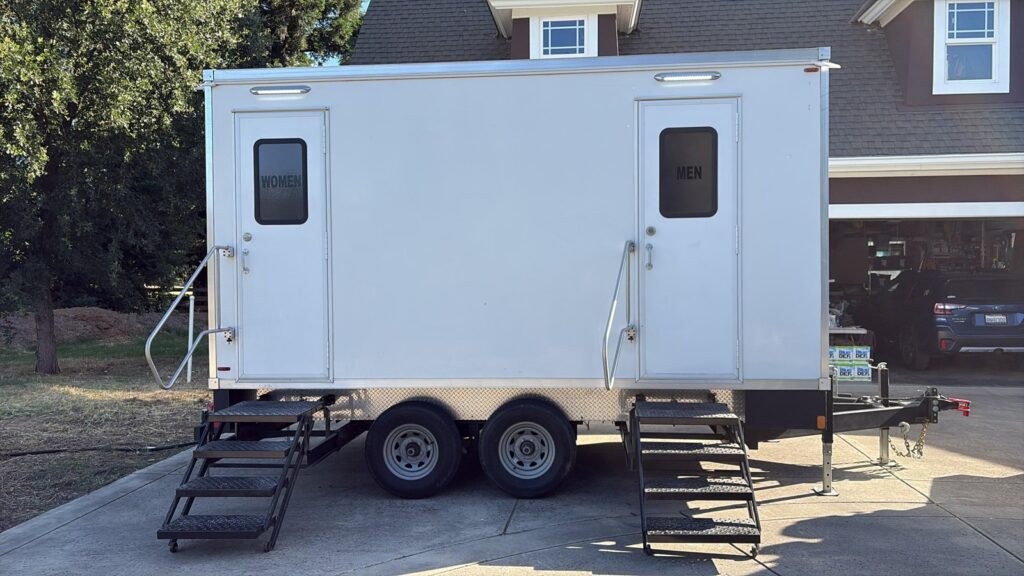 Side view of a portable bathroom trailer with women's and men's doors parked in a home driveway beside a two-car garage, The Lavatory Los Angeles