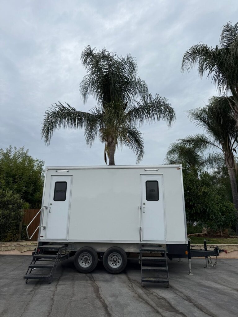 Portable bathroom trailer with women's and men's doors on an asphalt pad under tall palm trees, The Lavatory Los Angeles