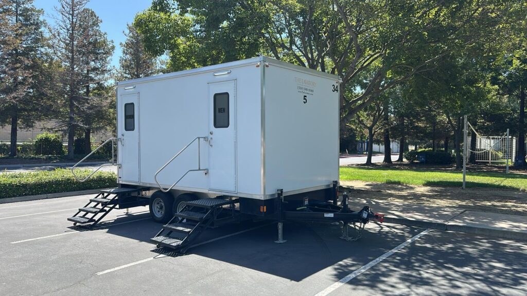 5-station bathroom trailer with women's and men's steps in a shaded parking lot next to a park lawn, The Lavatory Los Angeles