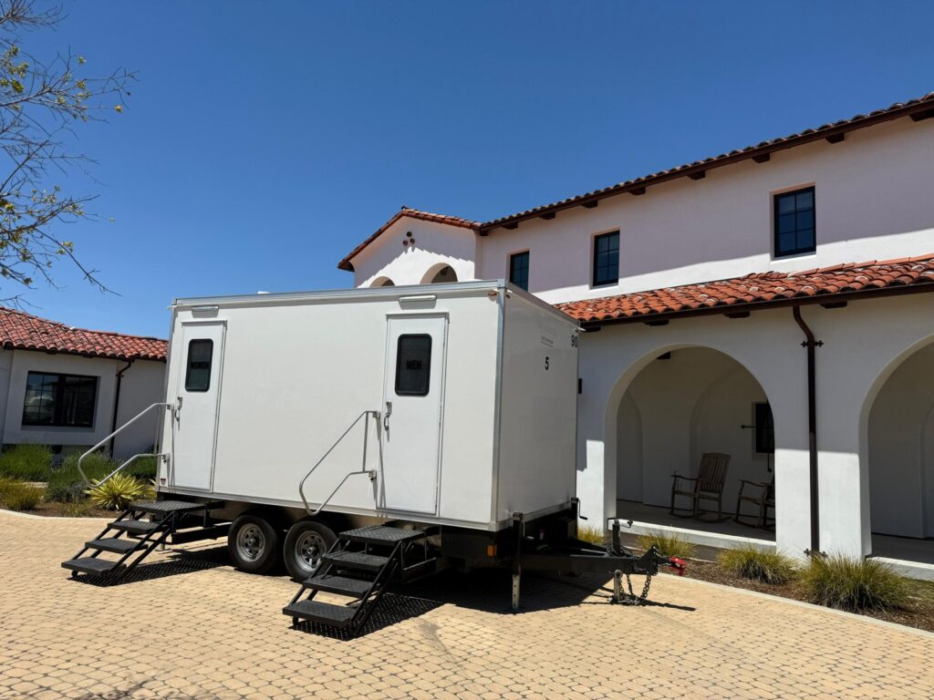 5-station luxury bathroom trailer with women's and men's doors and fold-down steps on a paver courtyard beside a white stucco building with a red tile roof, The Lavatory Los Angeles