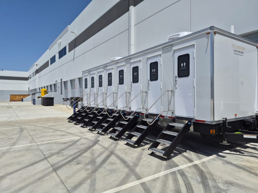 8-station private bathroom trailer with eight unisex stall doors and steps lined up along a new concrete warehouse, The Lavatory Los Angeles