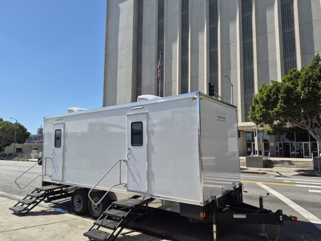 9-station bathroom trailer parked at the curb in front of a tall government building, The Lavatory Los Angeles