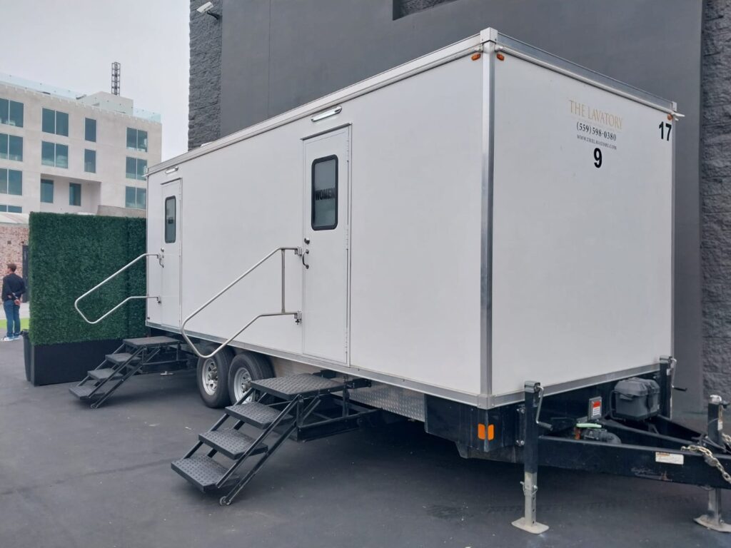 A member of the event staff walks past a 9-station bathroom trailer staged beside a hedge wall at a hotel event, The Lavatory Los Angeles