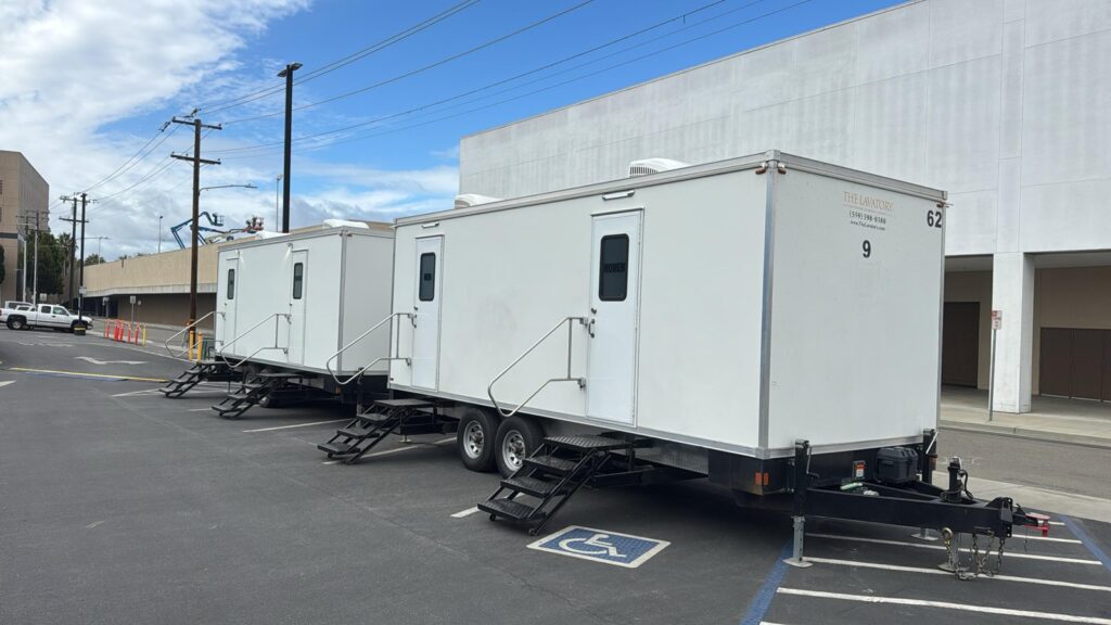 Two 9-station bathroom trailers parked end to end in a commercial parking lot under power lines, The Lavatory Los Angeles
