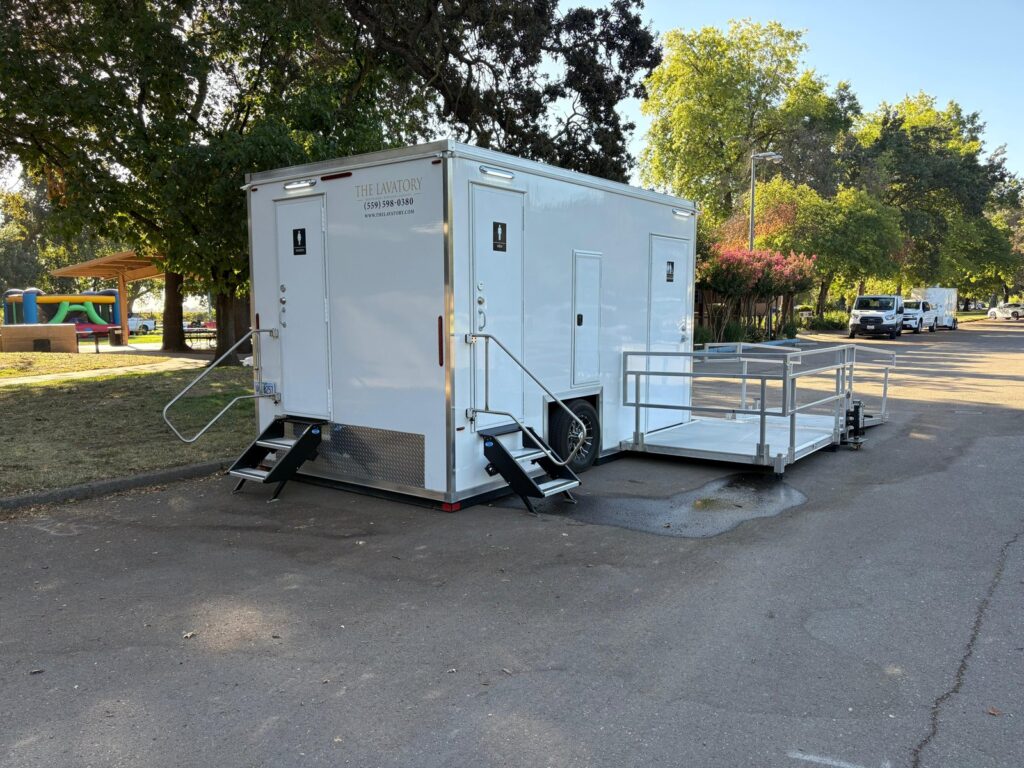 ADA plus 2 bathroom trailer with a railed platform parked on a park lot near a picnic shelter and a bounce house, The Lavatory Los Angeles