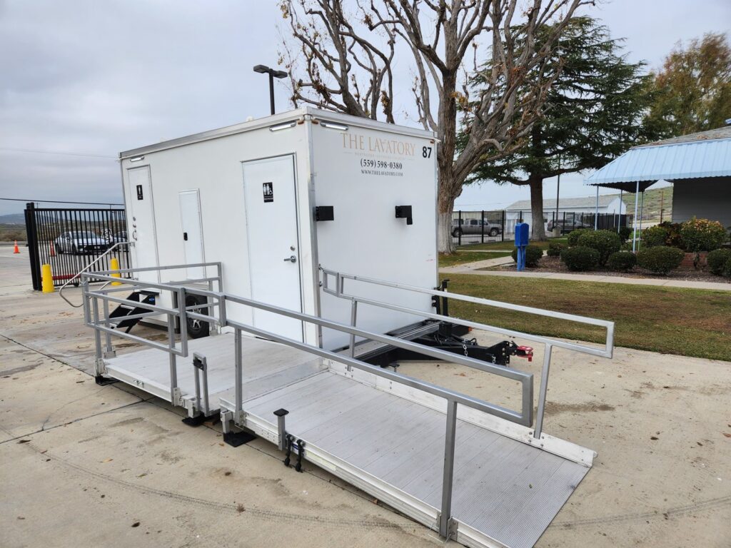 ADA plus 2 bathroom trailer with a long aluminum wheelchair ramp and railed platform on a concrete lot, The Lavatory Los Angeles