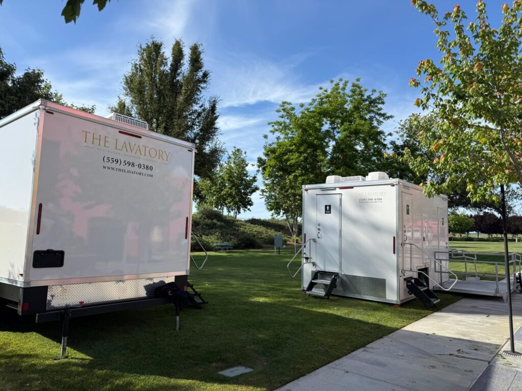 Two bathroom trailers, one with an ADA ramp platform, set on a park lawn beside a walkway, The Lavatory Los Angeles