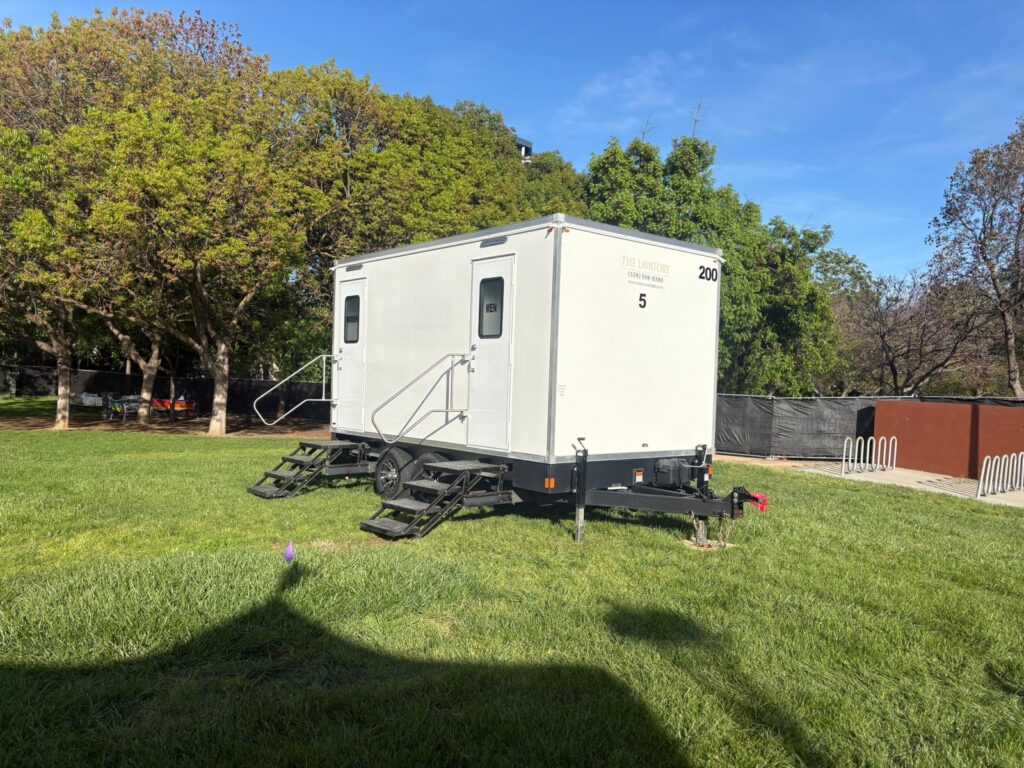 5-station bathroom trailer with women's and men's doors set on a park lawn under shade trees, The Lavatory Los Angeles
