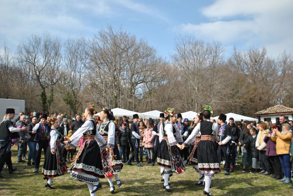A folk dance team in traditional dress circle dancing as a crowd of people watches at an outdoor festival, The Lavatory Los Angeles
