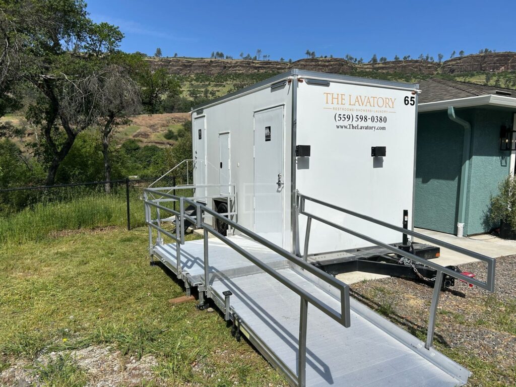 Accessible bathroom trailer with a long railed ramp set on a grassy hillside next to a home, The Lavatory Los Angeles