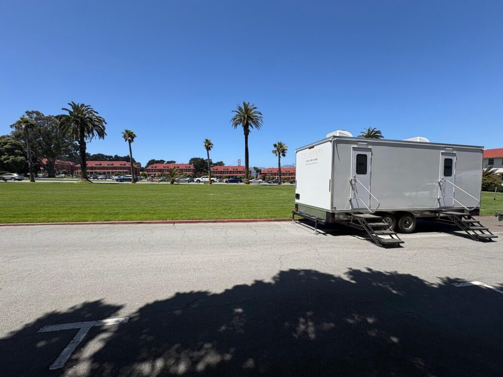 9-station bathroom trailer with two entry stairways parked on a paved lot beside a palm-lined lawn, The Lavatory Los Angeles