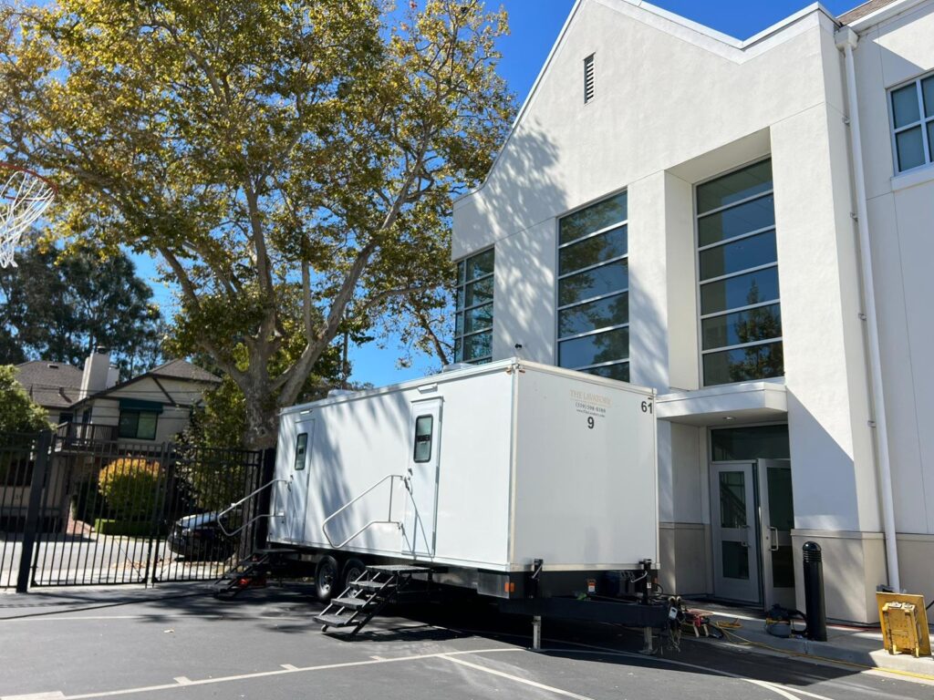 9-station bathroom trailer parked beside a white school building under shade trees, The Lavatory Los Angeles