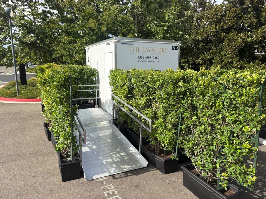 Accessible bathroom trailer with an aluminum ramp framed by potted hedge walls in a parking lot, The Lavatory Los Angeles