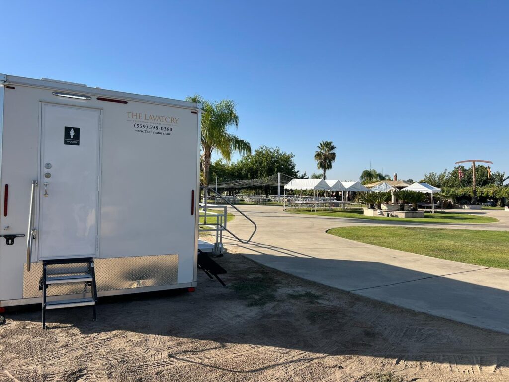 Bathroom trailer women's door and steps beside a curved drive, palm trees and white event tents, The Lavatory Los Angeles