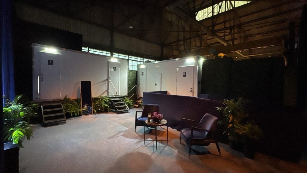 Luxury bathroom trailers set up inside a warehouse event space beside a lounge with chairs and plants, The Lavatory Los Angeles