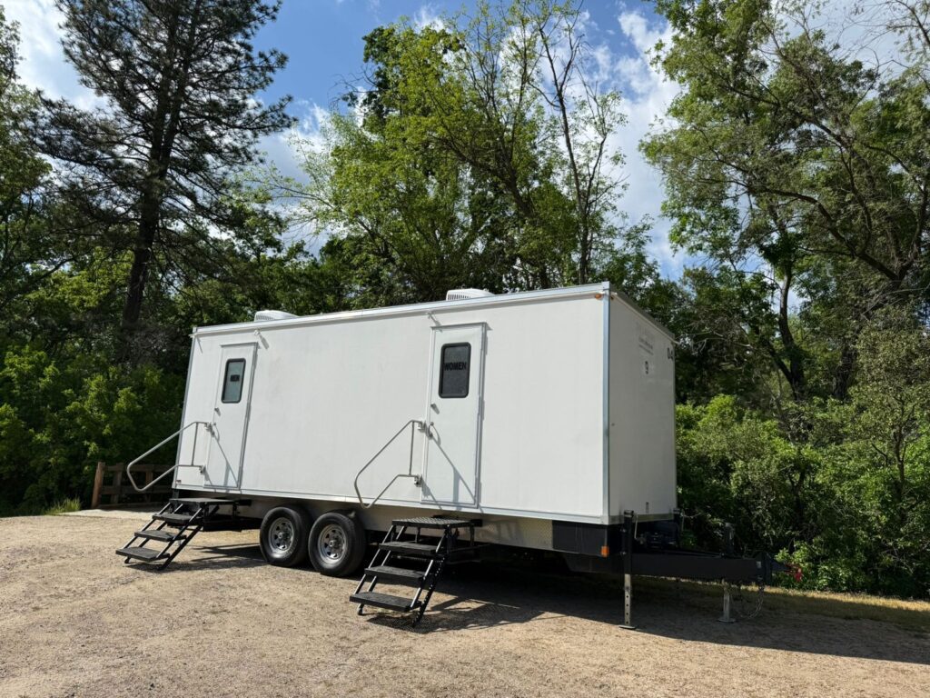 9-station bathroom trailer with two entrances set up on gravel at the edge of a wooded park, The Lavatory Los Angeles