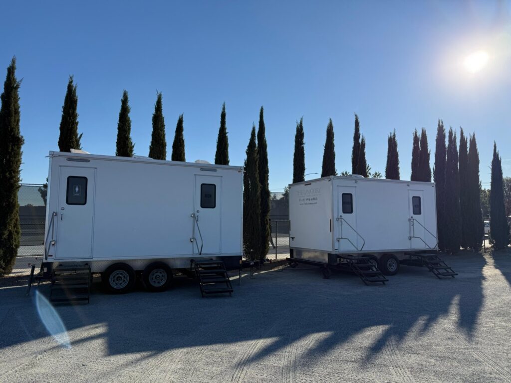 Two bathroom trailers parked side by side in a lot lined with tall cypress trees, The Lavatory Los Angeles
