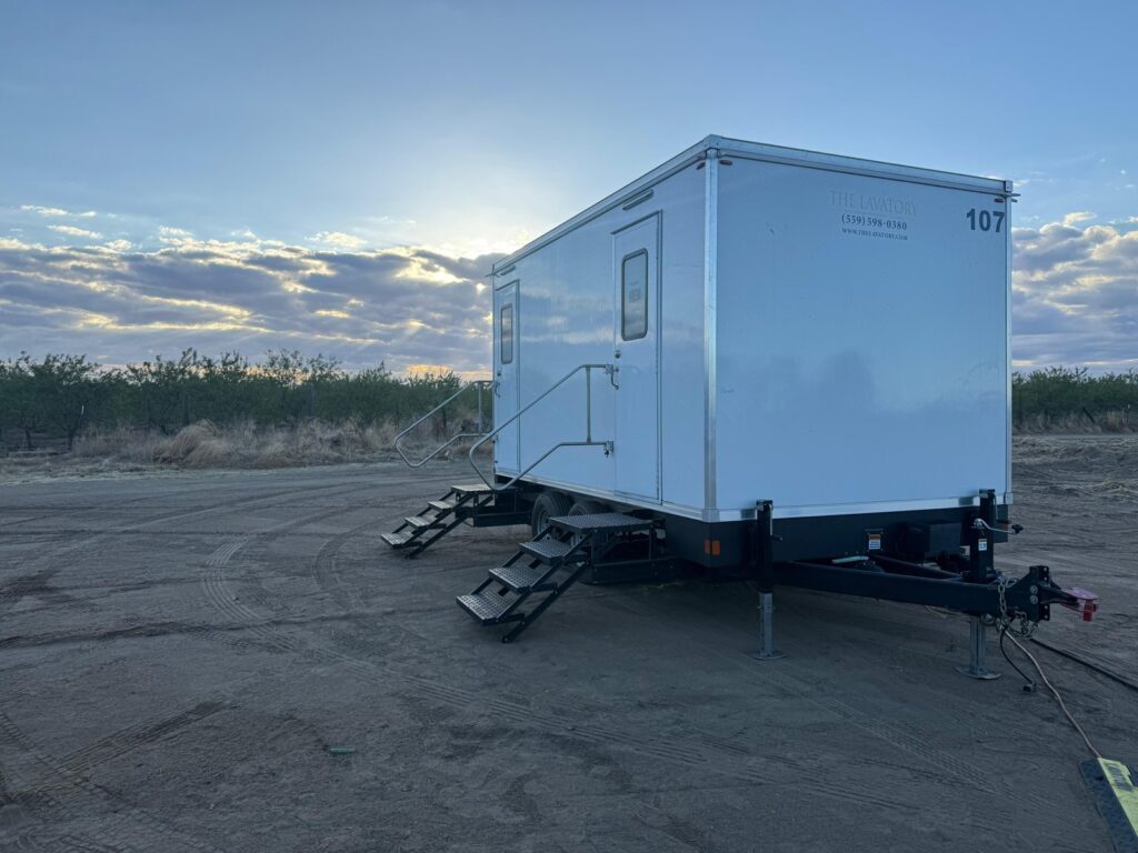 2-station bathroom trailer parked on a packed dirt lot at dusk with the sun low behind it, The Lavatory Los Angeles