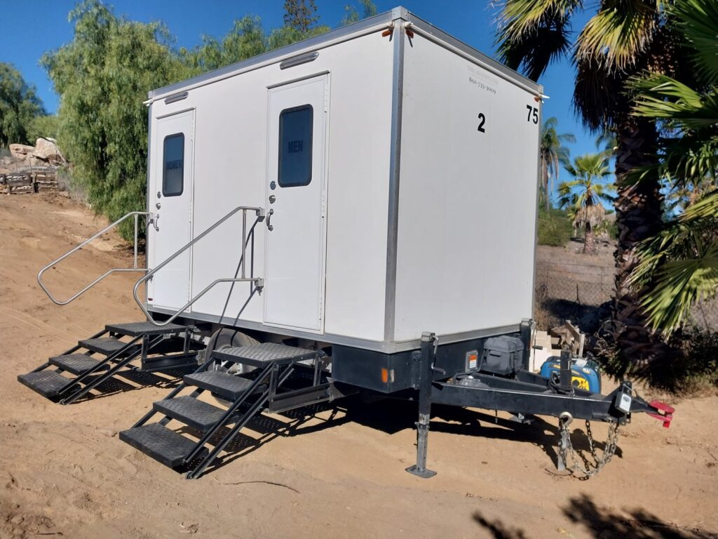 2-station bathroom trailer with fold-down steps set on a sandy desert lot beside palms and boulders, The Lavatory Los Angeles
