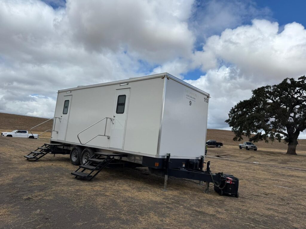 9-station bathroom trailer with steps, handrails and a generator on dry golden grassland under big clouds, The Lavatory Los Angeles