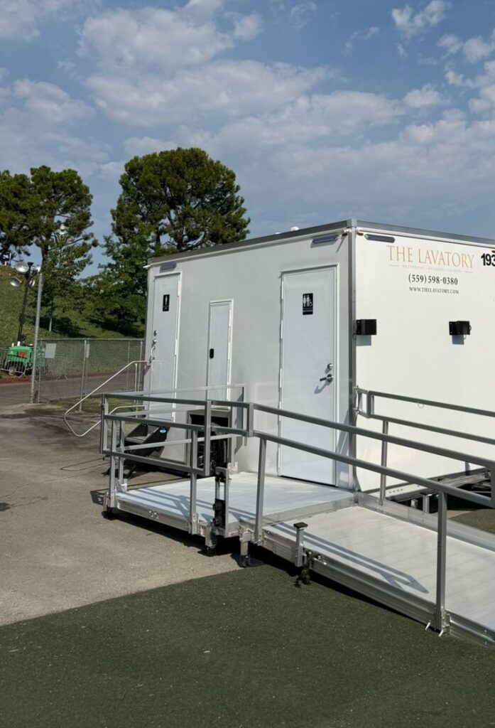 Accessible bathroom trailer with a railed wheelchair ramp on turf beside a ball field, The Lavatory Los Angeles