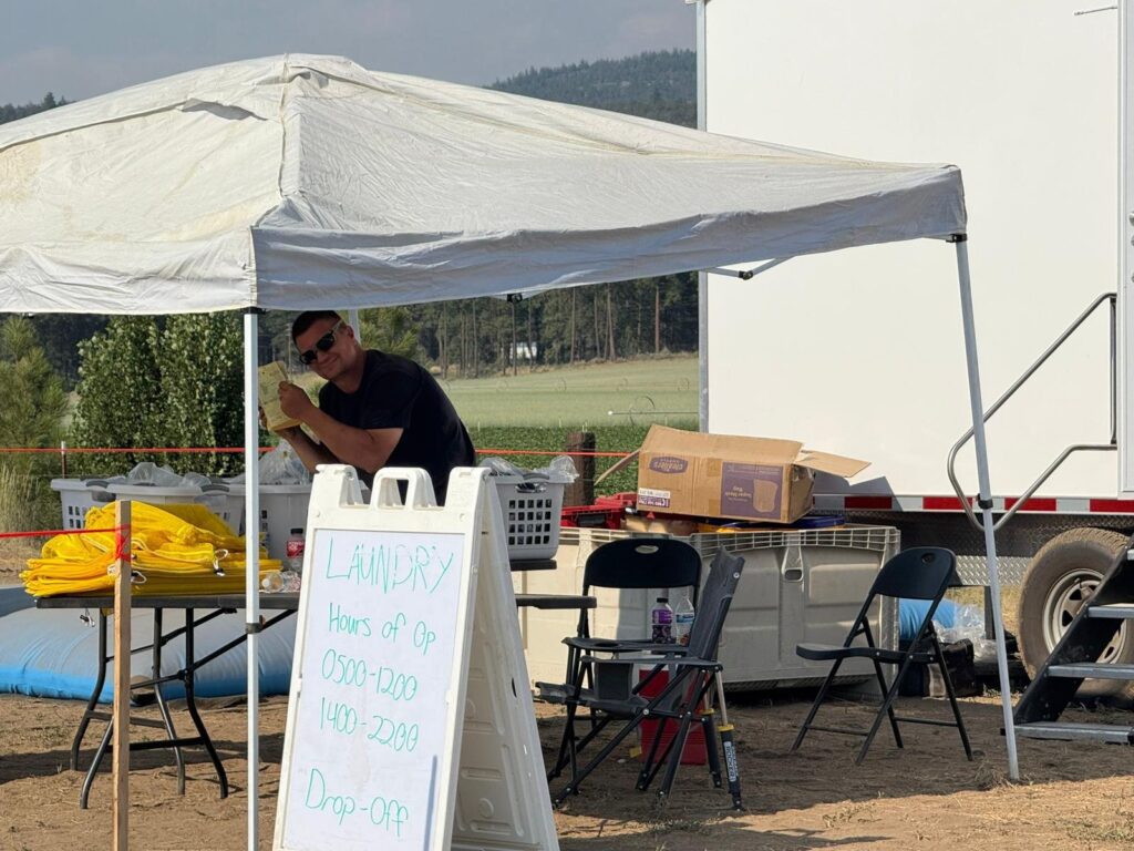 Laundry staff member at a drop-off tent with an hours sign beside a mobile laundry trailer, The Lavatory Los Angeles