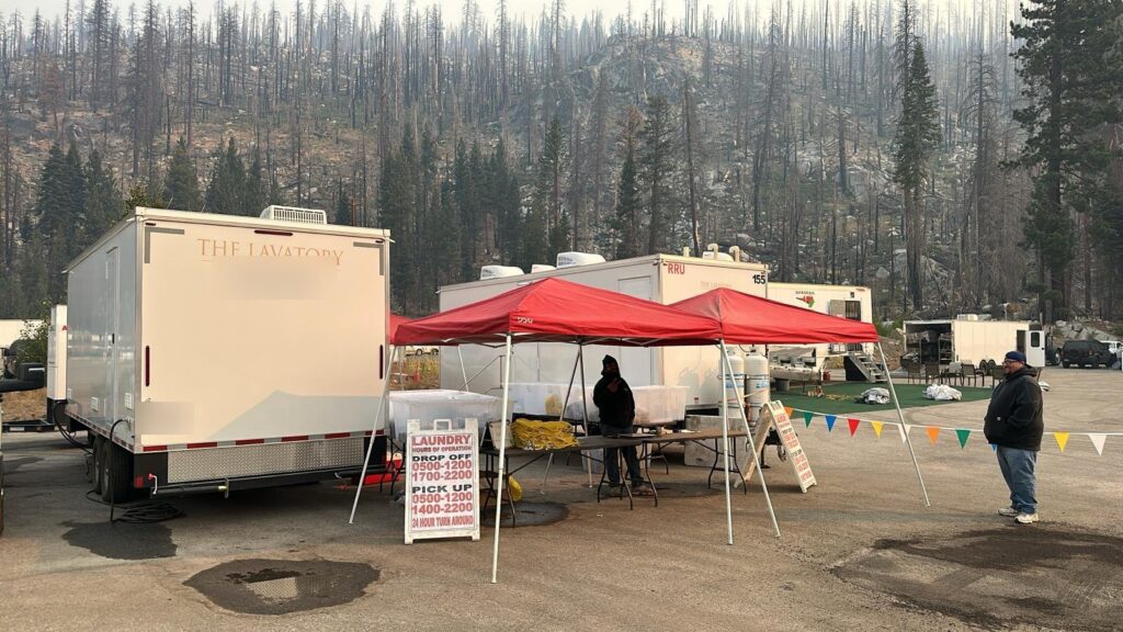 People at a laundry drop-off canopy beside a laundry trailer at a fire camp below a burned forest, The Lavatory Los Angeles