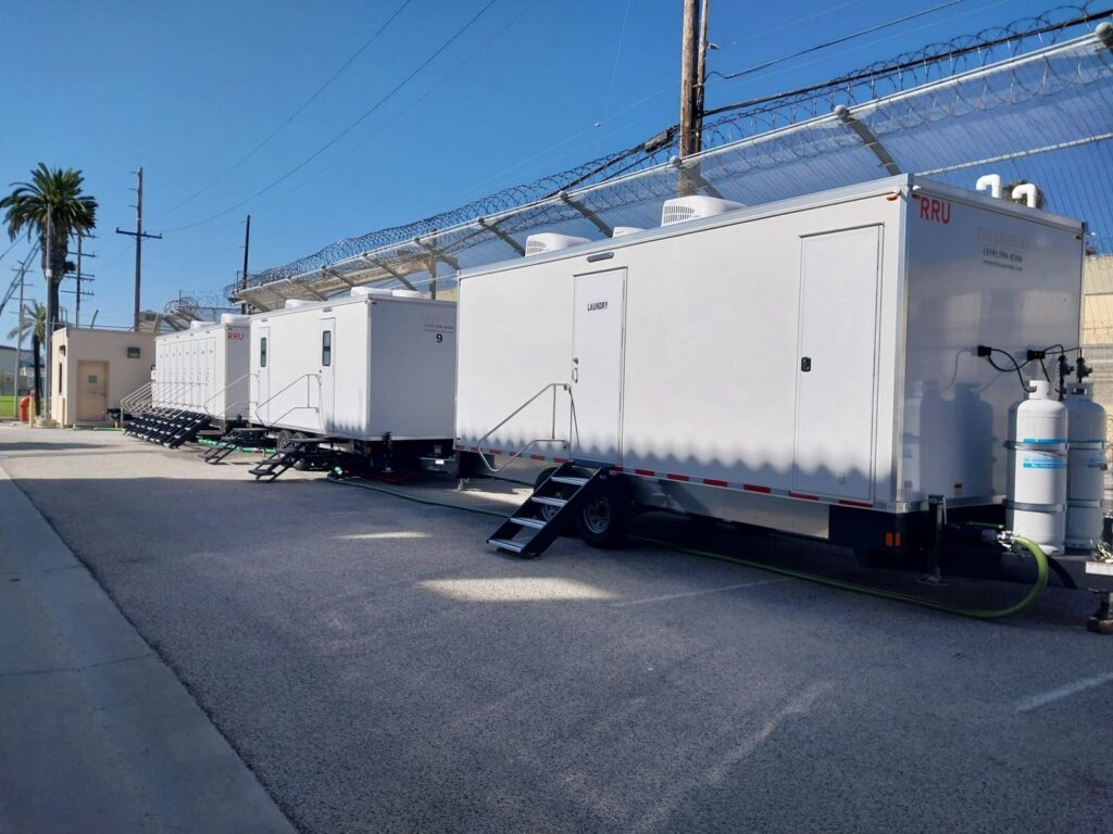 Mobile laundry trailer lined up with bathroom trailers along a fenced lot with palm trees, The Lavatory Los Angeles