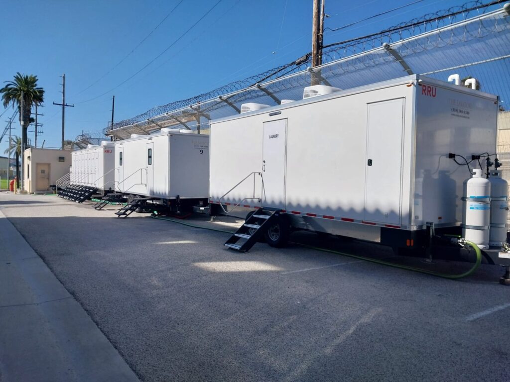 Mobile laundry trailer at the head of a row of restroom and shower trailers along a fenced lot with palm trees, The Lavatory Los Angeles