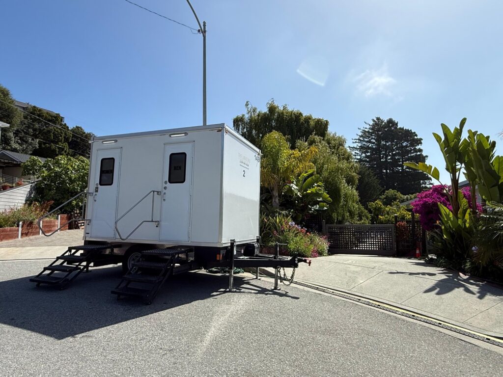 2-station bathroom trailer with fold-down steps parked on a sloped residential street beside a garden, The Lavatory Los Angeles