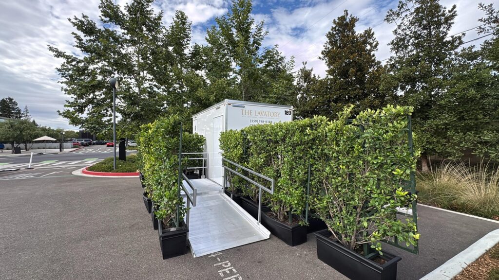 Accessible bathroom trailer with an aluminum ramp framed by hedge planters in a paved lot, The Lavatory Los Angeles