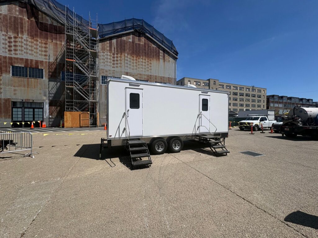 Luxury bathroom trailer with two entry doors parked beside a historic industrial building wrapped in scaffolding, The Lavatory Los Angeles