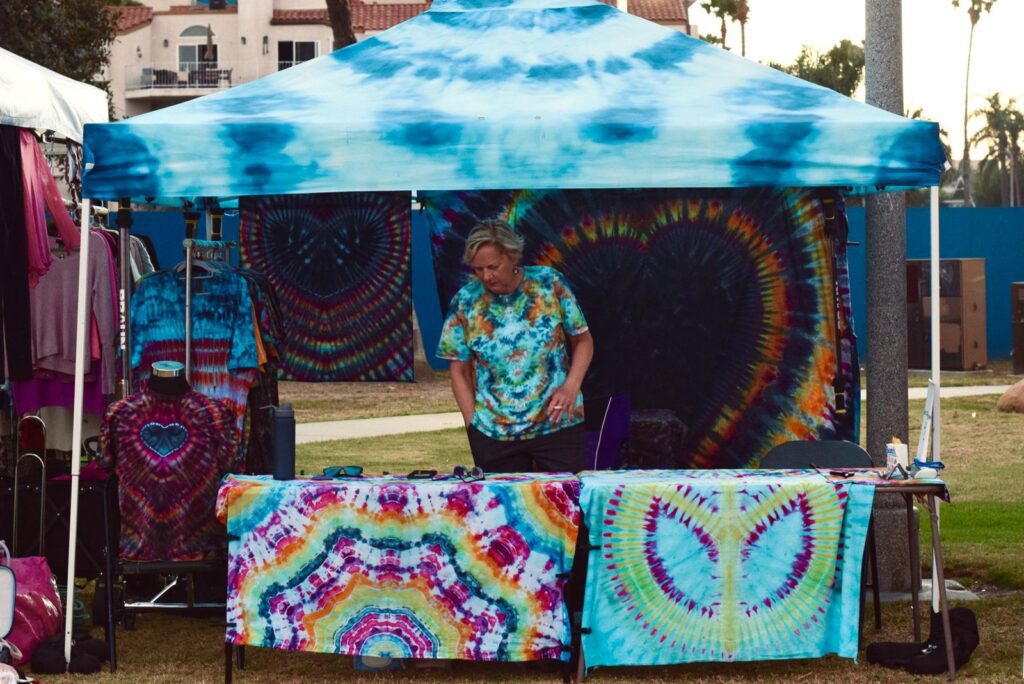 Festival vendor staff arranging tie-dye shirts under a canopy booth at an outdoor street fair, The Lavatory Los Angeles
