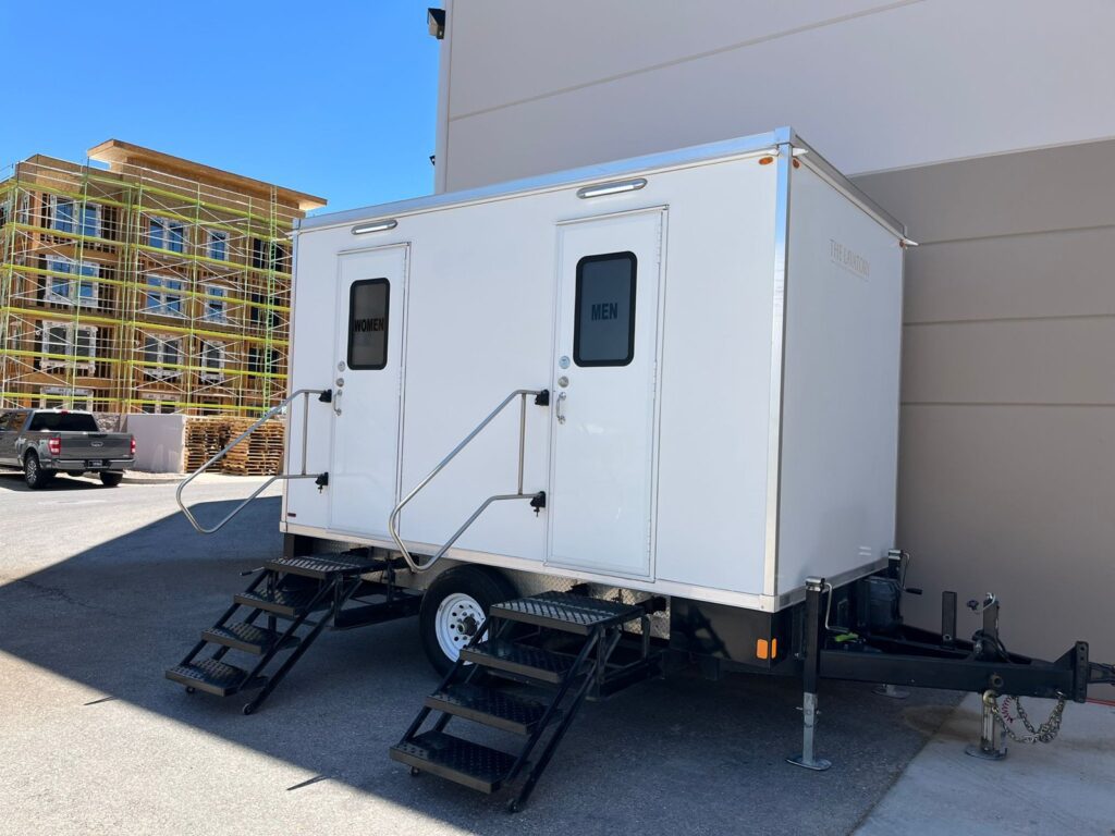 2-station bathroom trailer with women's and men's doors beside an apartment building under construction, The Lavatory Los Angeles