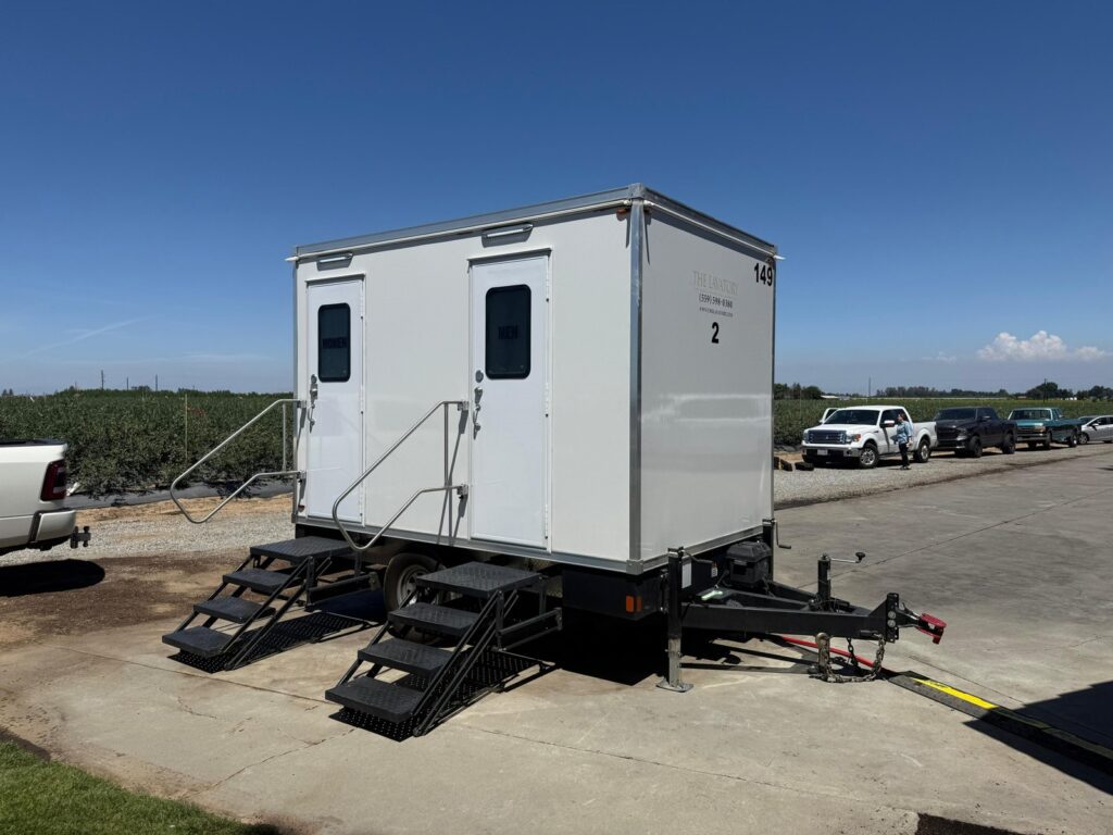 2-station bathroom trailer on a concrete pad next to a berry field, with farm workers and their trucks parked nearby, The Lavatory Los Angeles