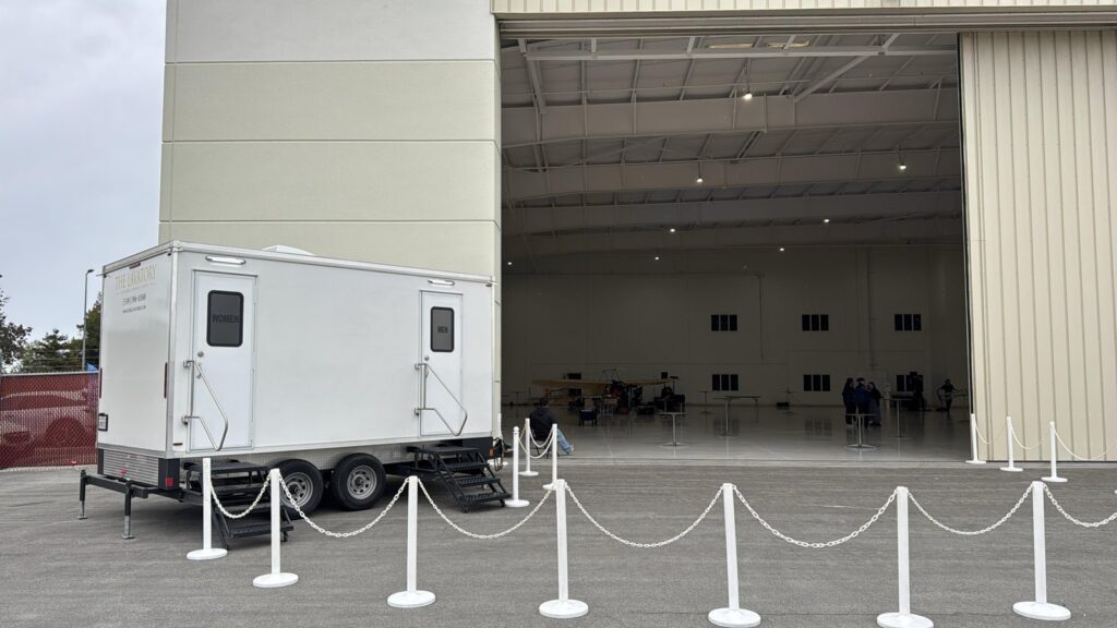 Bathroom trailer with women's and men's doors set outside an open aircraft hangar behind rope stanchions, The Lavatory Los Angeles
