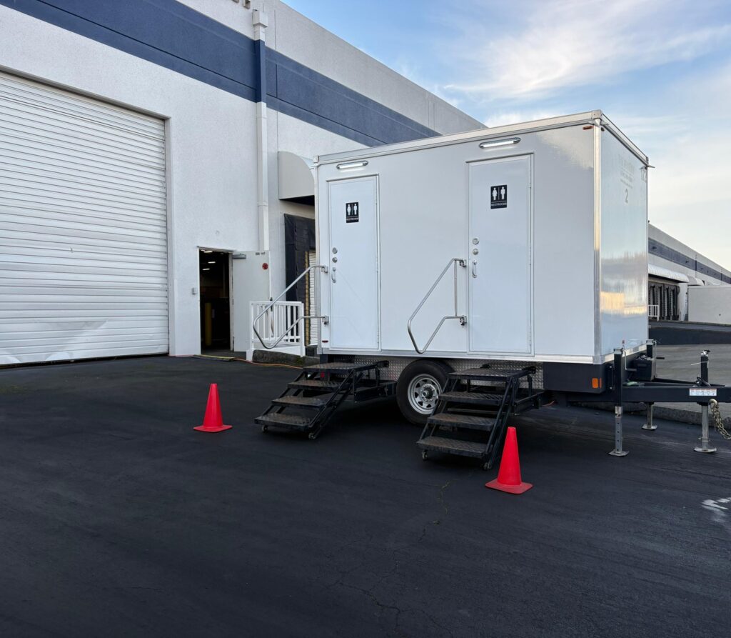 2-station bathroom trailer with safety cones beside a roll-up door at an industrial building, The Lavatory Los Angeles