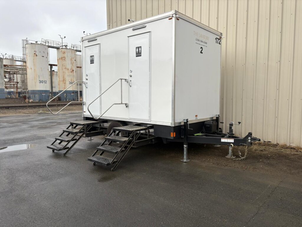 2-station bathroom trailer parked against a metal building with storage tanks at a processing plant behind it, The Lavatory Los Angeles