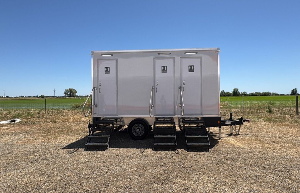 3-station bathroom trailer with three doors and steps parked on dry ground at the edge of a green crop field, The Lavatory Los Angeles