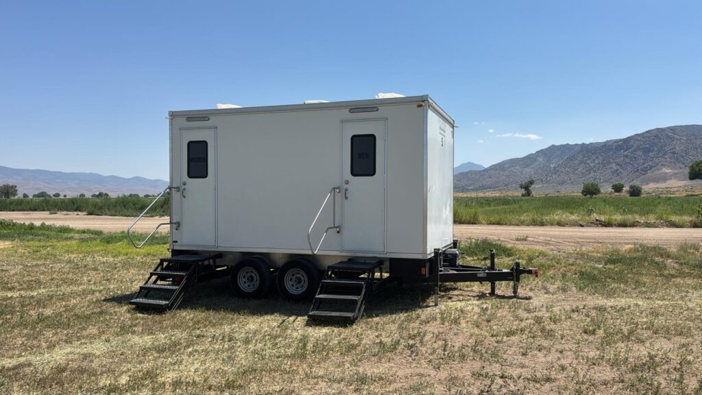 Portable bathroom trailer with two entry doors and steps on an open field below brown mountains, The Lavatory Los Angeles